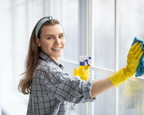 Smiling young woman with detergent and rag cleaning window glass. Professional sanitary service